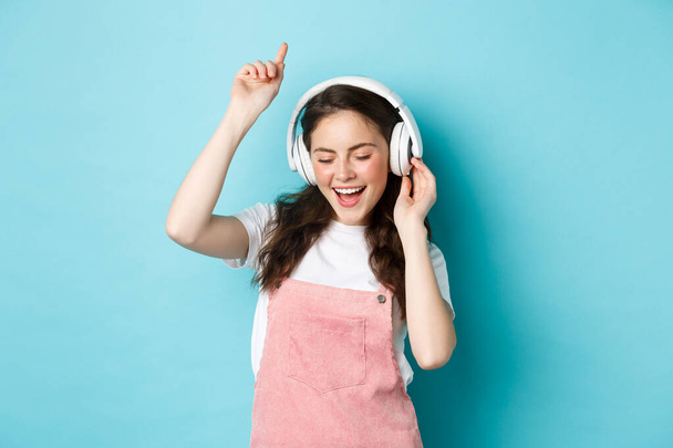 Κομψό κορίτσι της χιλιετίας που χορεύει με ανοιξιάτικο ντύσιμο, ακούει μουσική με ακουστικά και απολαμβάνει τον ήχο, στέκεται πάνω από το μπλε φόντο - Φωτογραφία, εικόνα