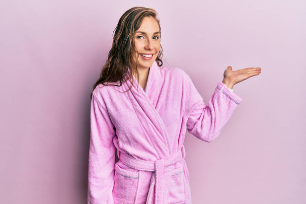 Young blonde woman wearing bathrobe smiling cheerful presenting and pointing with palm of hand looking at the camera.  - Foto, Imagen