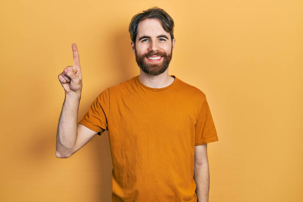 Caucasian man with beard wearing casual yellow t shirt showing and pointing up with finger number one while smiling confident and happy.  - Photo, Image