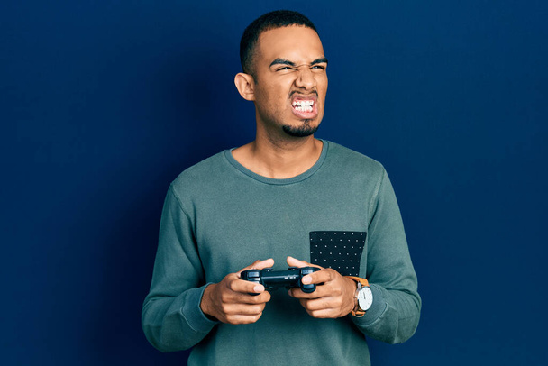 Young african american man playing video game holding controller angry and mad screaming frustrated and furious, shouting with anger. rage and aggressive concept.  - Photo, Image