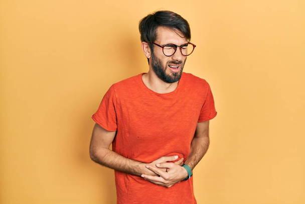 Young hispanic man wearing casual clothes and glasses with hand on stomach because nausea, painful disease feeling unwell. ache concept.  - Foto, afbeelding