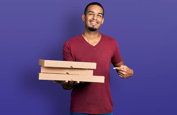 Joven afroamericano hombre sosteniendo llevar comida sonriendo feliz señalando con la mano y el dedo  - Foto, imagen