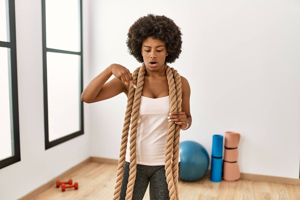 Νεαρή Αφροαμερικανή γυναίκα με αφρο μαλλιά στο γυμναστήριο προπόνηση με σχοινιά μάχης που δείχνουν προς τα κάτω με δάχτυλα που δείχνουν διαφήμιση, έκπληκτη φάτσα και ανοιχτό στόμα  - Φωτογραφία, εικόνα