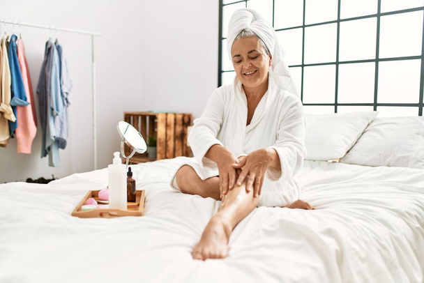 Grauhaarige Frau mittleren Alters pflegt die Beine der Haut mit Feuchtigkeitscreme, die auf dem Bett im Schlafzimmer sitzt. - Foto, Bild