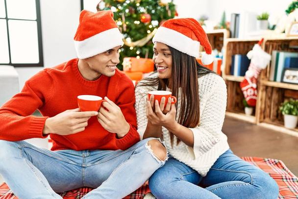 Młoda Latynoska para uśmiechnięta szczęśliwa pijąca kawę siedząca na podłodze przy choince w domu. - Zdjęcie, obraz