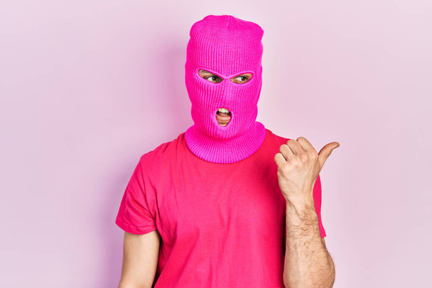 Молодой латиноамериканец с современными окрашенными волосами, одетый в розовую маску балаклавы, указывает пальцем вверх в сторону улыбаясь счастливо с открытым ртом  - Фото, изображение