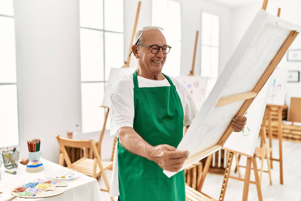 starszy siwowłosy artysta człowiek uśmiecha szczęśliwy trzymając płótno w studio sztuki. - Zdjęcie, obraz