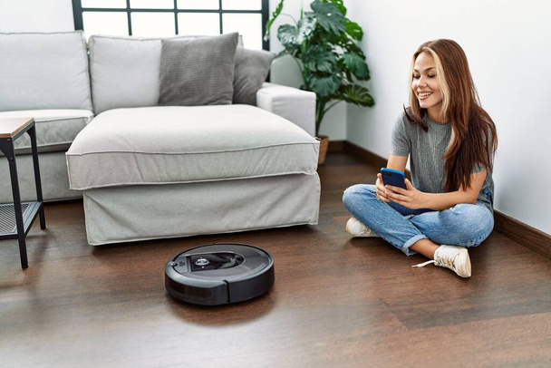 Young caucasian girl controlling robot vacuum cleaner with smartphone sitting on the floor at home. - Photo, Image