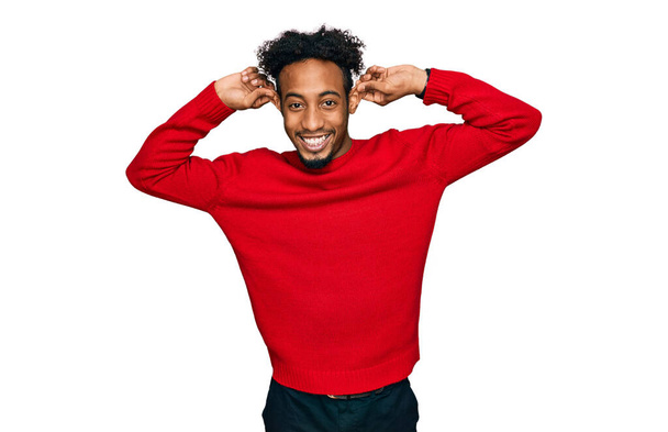 Jovem afro-americano com barba vestindo camisola de inverno casual sorrindo puxando orelhas com os dedos, gesto engraçado. problema de audição  - Foto, Imagem