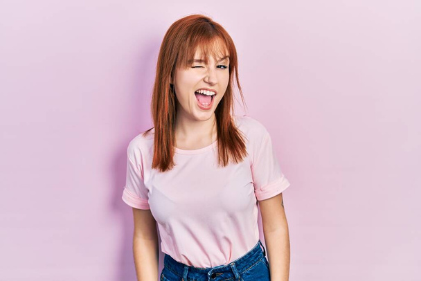 Рыжая молодая женщина в обычной розовой футболке подмигивает, глядя в камеру с сексуальным выражением лица, веселым и счастливым лицом.  - Фото, изображение