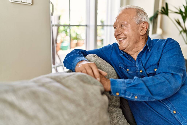 Vanhempi mies, jolla on harmaat hiukset, istuu sohvalla talonsa olohuoneessa. Kypsä mies hymyilee onnellinen kotona. - Valokuva, kuva