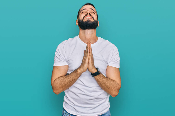 Young man with beard wearing casual white t shirt begging and praying with hands together with hope expression on face very emotional and worried. begging.  - Фото, изображение