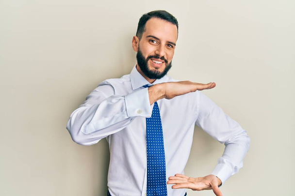 Молодой человек с бородой носит деловой галстук жест с руками показывая большой и большой знак размера, мера символа. улыбаясь, глядя в камеру. концепция измерения.  - Фото, изображение