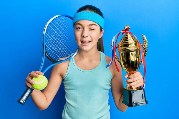 Bella bruna bambina che gioca a tennis con trofeo in mano ammiccando guardando la fotocamera con espressione sexy, viso allegro e felice.  - Foto, immagini