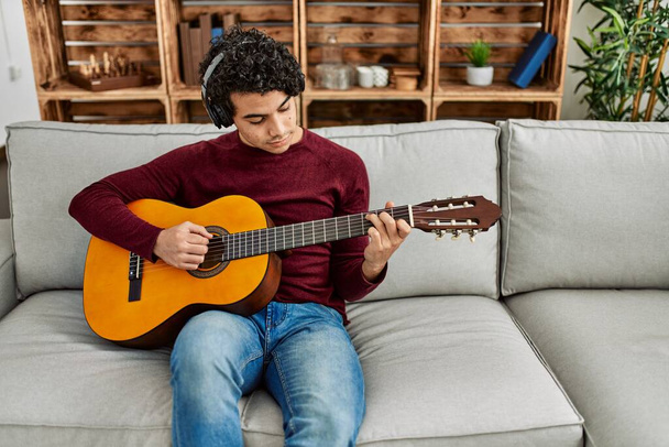 Jovem hispânico tocando guitarra clássica sentado no sofá em casa. - Foto, Imagem