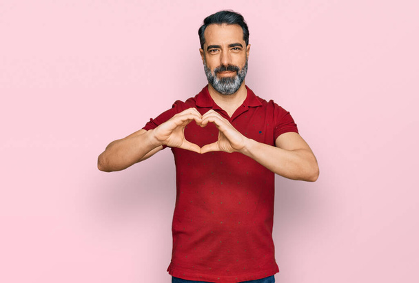 Man van middelbare leeftijd met baard in casual rood t-shirt glimlachend in liefde met hartsymbool en vorm met handen. romantisch concept.  - Foto, afbeelding