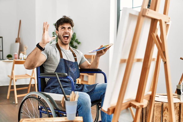 Νεαρός Ισπανόφωνος κάθεται σε αναπηρική καρέκλα ζωγραφική στο στούντιο τέχνης τρελός και τρελός φωνάζοντας και φωνάζοντας με επιθετική έκφραση και τα χέρια ψηλά. έννοια απογοήτευσης.  - Φωτογραφία, εικόνα