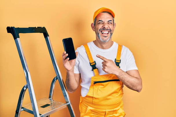 Guapo hombre de mediana edad con el pelo gris de pie por escalera que muestra el teléfono inteligente sonriendo y riendo en voz alta porque broma loca divertida.  - Foto, Imagen