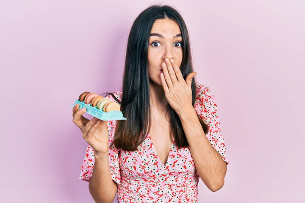 Junge brünette Frau mit köstlichen Macarons Gebäck den Mund mit der Hand bedeckt, schockiert und Angst vor Fehler. Überraschte Miene  - Foto, Bild