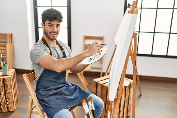 Joven artista hispano sonriendo feliz pintura en el estudio de arte. - Foto, Imagen