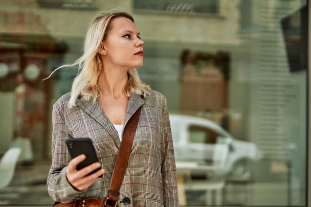Young blonde businesswoman with serious expression using smartphone at the city. - Фото, зображення
