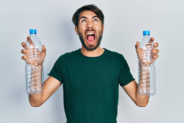 Joven hispano sosteniendo botellas de plástico de reciclaje enojado y loco gritando frustrado y furioso, gritando con ira mirando hacia arriba.  - Foto, Imagen