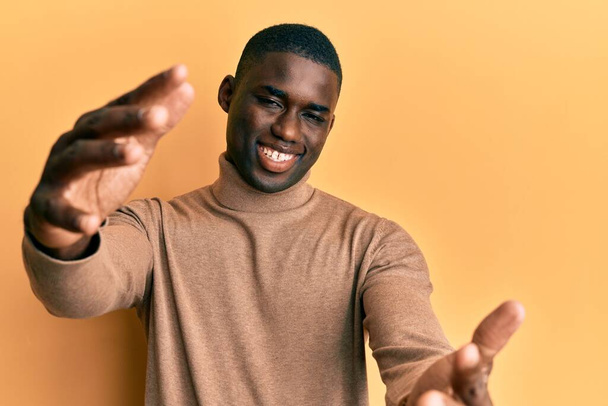 Junger afrikanisch-amerikanischer Mann in lässigem Winterpullover, der lächelnd mit offenen Armen in die Kamera blickt, um sich zu umarmen. fröhlicher Ausdruck, der das Glück umarmt.  - Foto, Bild