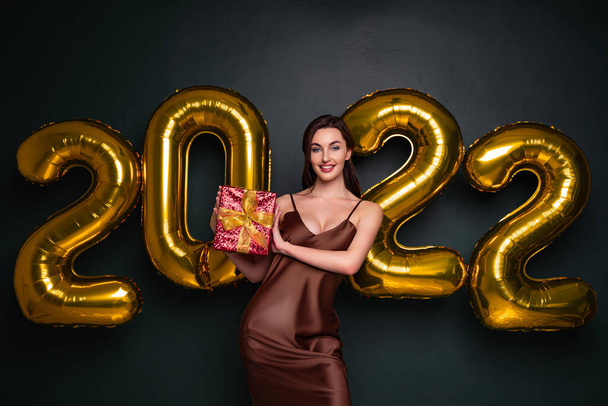 Georgeous brunette lady in beautiful dress holding gift box on black studio background with golden 2022 balloons, panorama. Happy young woman with wrapped present at New Year party - Фото, изображение
