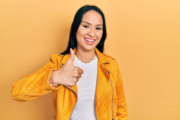 Mooie Latijns-Amerikaanse vrouw met neuspiercing in geel leren jasje doet vrolijke duimen omhoog gebaar met de hand. goedkeuring van expressie kijken naar de camera toont succes.  - Foto, afbeelding