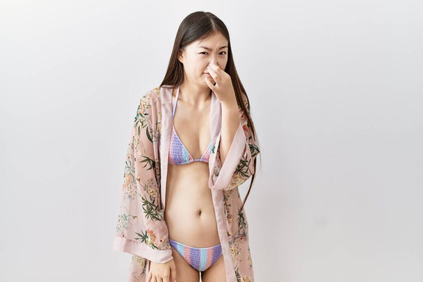 Mujer asiática joven usando bikini un kimono floral oliendo algo apestoso y asqueroso, olor intolerable, conteniendo la respiración con los dedos en la nariz. mal olor  - Foto, Imagen
