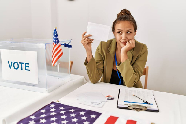 Beautiful hispanic woman holding voting envelop in ballot box serious face thinking about question with hand on chin, thoughtful about confusing idea  - Photo, Image
