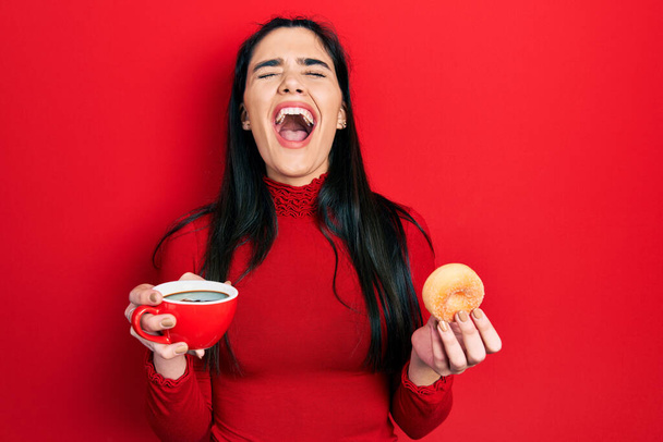 Jong Spaans meisje drinken van koffie en het eten van gebak boos en gek schreeuwen gefrustreerd en woedend, schreeuwen van woede opkijken.  - Foto, afbeelding