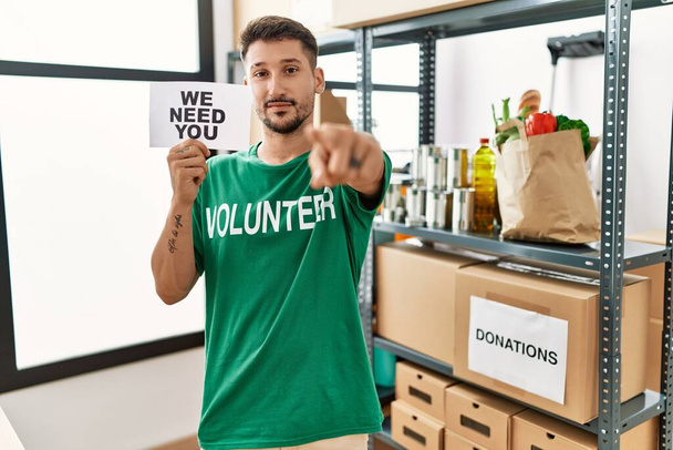 Młody Latynos noszący koszulkę ochotnika, trzymający sztandar wskazujący palcem na kamerę i na ciebie, pewny siebie gest wyglądający poważnie.  - Zdjęcie, obraz