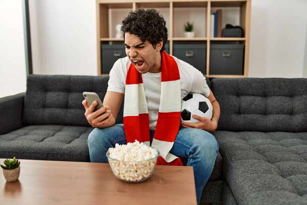 Jonge Spaanse man kijkt naar voetbalwedstrijd en ondersteunend team thuis. - Foto, afbeelding