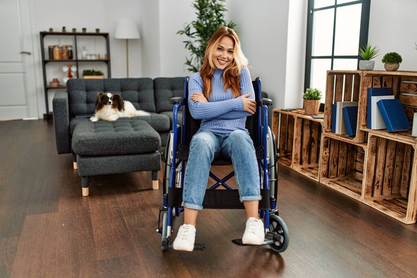 Νεαρή όμορφη γυναίκα κάθεται σε αναπηρική καρέκλα στο σπίτι χαρούμενο πρόσωπο χαμογελώντας με σταυρωμένα χέρια κοιτάζοντας την κάμερα. θετικό πρόσωπο.  - Φωτογραφία, εικόνα