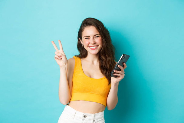 Technology and lifestyle concept. Beautiful cheerful girl sending positive vibes, smiling and showing peace sign, using cellphone, standing against blue background - Φωτογραφία, εικόνα
