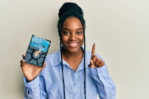 Femme afro-américaine aux cheveux tressés tenant le disque dur souriant avec une idée ou une question pointant du doigt avec un visage heureux, numéro un  - Photo, image