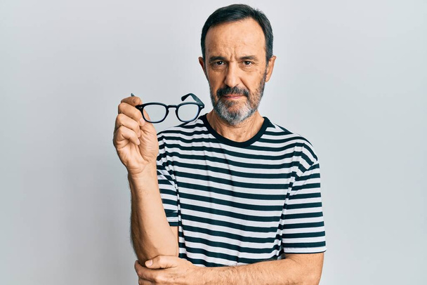 Middelbare leeftijd Spaanse man het dragen van casual kleding en het houden van een bril ontspannen met ernstige uitdrukking op het gezicht. eenvoudig en natuurlijk kijkend naar de camera.  - Foto, afbeelding