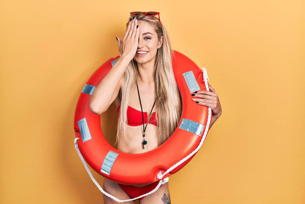 Mujer caucásica joven que usa bikini y sostiene salvavidas flotando cubriendo un ojo con la mano, sonrisa segura en la cara y emoción sorpresa.  - Foto, Imagen