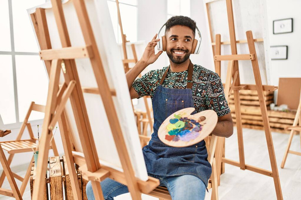młody afrykański artysta amerykański człowiek uśmiechnięty szczęśliwy słuchając muzyki i malarstwo w studio sztuki. - Zdjęcie, obraz
