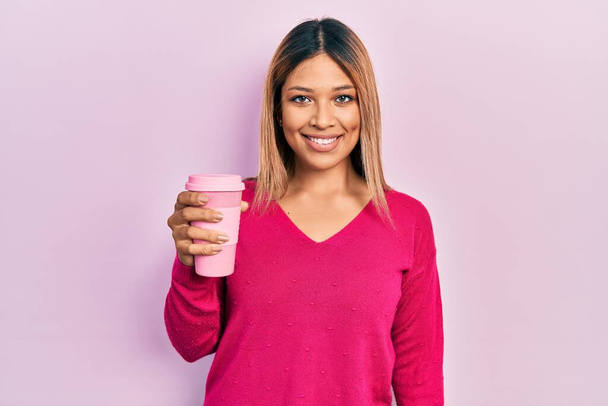 Piękna Latynoska kobieta pije filiżankę kawy patrząc pozytywnie i szczęśliwie stojąc i uśmiechając się z ufnym uśmiechem pokazując zęby  - Zdjęcie, obraz