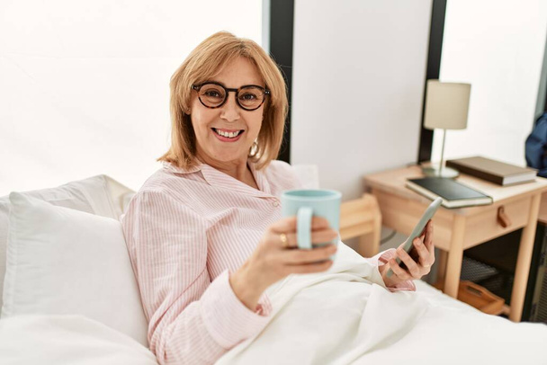 Блондинка середнього віку лежить на ліжку, використовуючи смартфон і п'є каву вдома
. - Фото, зображення