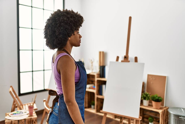 Mujer afroamericana joven con pelo afro en el estudio de arte mirando a un lado, pose de perfil relajante con rostro natural y sonrisa confiada.  - Foto, Imagen