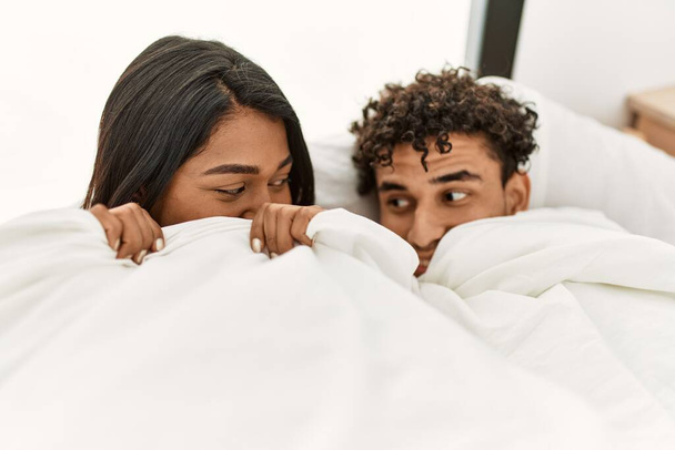 Mladý latinský pár krytí s prostěradlem ležící na posteli v ložnici. - Fotografie, Obrázek