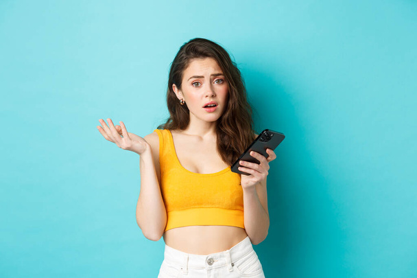 Technology and lifestyle concept. Glamour girl looks confused after reading smartphone screen, staring puzzled at camera, standing against blue background - Fotó, kép