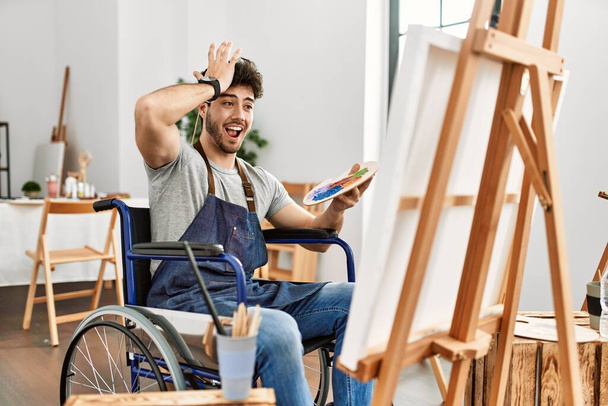 Jeune homme hispanique assis sur la peinture en fauteuil roulant au studio d'art surpris avec la main sur la tête pour erreur, rappelez-vous erreur. oublié, mauvais concept de mémoire.  - Photo, image