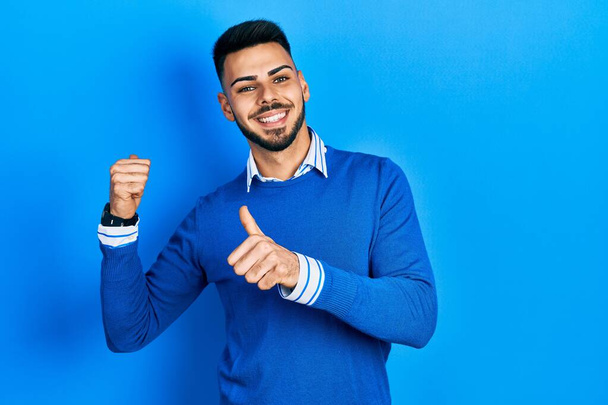 Mladý hispánec s plnovousem na sobě nenucený modrý svetr ukazující dozadu s rukou a palci nahoru, s úsměvem sebevědomý  - Fotografie, Obrázek