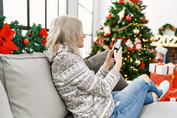 Nuori valkoihoinen tyttö istuu sohvalla älypuhelimen avulla kotona. - Valokuva, kuva