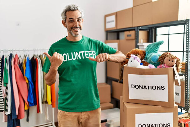 Латиноамериканец средних лет, одетый в футболку добровольца на пожертвования, указывает на спину с поднятой рукой и большим пальцем, уверенно улыбаясь  - Фото, изображение