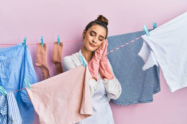 Beautiful brunette young woman washing clothes at clothesline sleeping tired dreaming and posing with hands together while smiling with closed eyes.  - Photo, Image